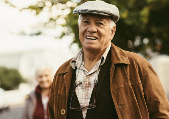 Portrait of happy senior man in warm clothes and cap looking at camera with a woman in background. Retired man standing outside on street on a winter day.